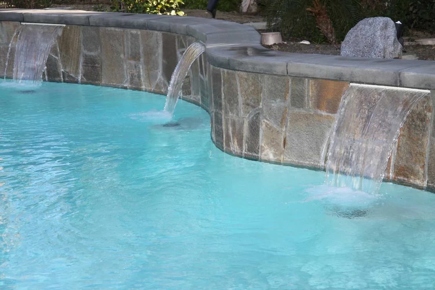 A swimming pool with three sheer-descent water features spilling over a flagstone wall